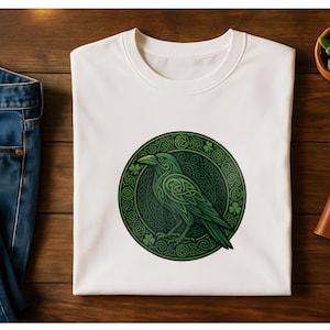 May include: White t-shirt featuring a green raven design within a circular Celtic knot pattern. The raven is detailed with intricate knotwork, surrounded by shamrocks. The shirt is folded on a wooden surface, next to blue jeans.