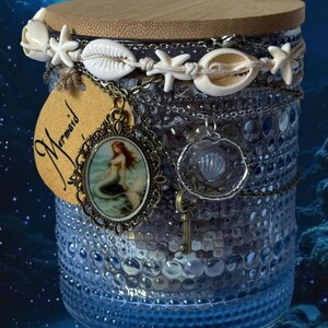 Op de afbeelding: Een blauwe glazen pot met een houten deksel, versierd met schelpen, zeesterren en kettingen. Een label met het woord "Mermaid" en een zeemeermin hanger hangen aan de pot. De pot heeft een gestructureerd oppervlak tegen een blauwe onderwater achtergrond.