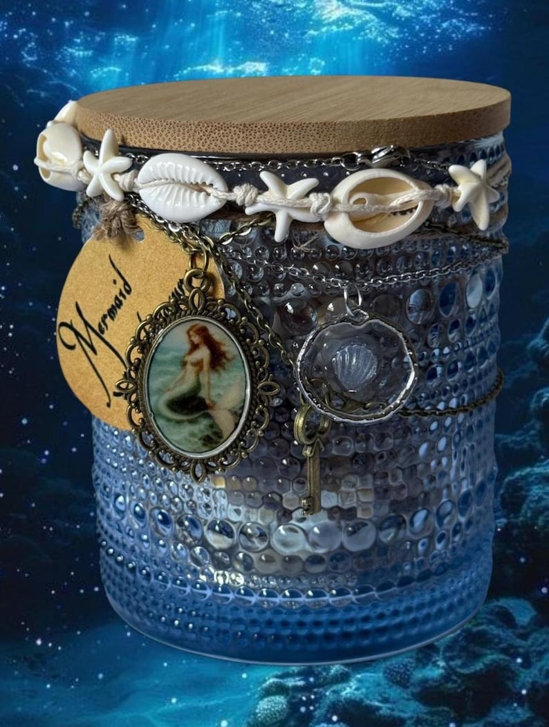 Op de afbeelding: Een blauwe glazen pot met een houten deksel, versierd met schelpen, zeesterren en kettingen. Een label met het woord "Mermaid" en een zeemeermin hanger hangen aan de pot. De pot heeft een gestructureerd oppervlak tegen een blauwe onderwater achtergrond.