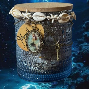Op de afbeelding: Een blauwe glazen pot met een houten deksel, versierd met schelpen en een zeemeermin hanger. De pot heeft een gestructureerd oppervlak en is versierd met kettingen en een sleutelbedel. Het woord "Mermaid" staat op een label.