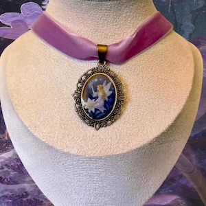 May include: A lavender velvet choker necklace with a bronze-colored pendant. The oval pendant features a fairy illustration with a white flower and a starry night sky. The necklace is displayed on a mannequin.