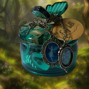 May include: A teal glass jar with a butterfly-shaped lid and two pendant necklaces. The pendants feature fairy silhouettes in oval frames. A tag reads "fairy bottle". The jar is decorated with leaf accents.