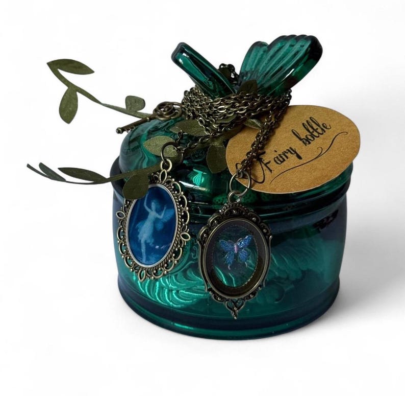 May include: A teal glass jar with a lid, adorned with a chain and two oval pendants. One pendant features a blue ballerina, the other a blue butterfly. A tag reads "Fairy bottle". Decorative green leaves are attached.