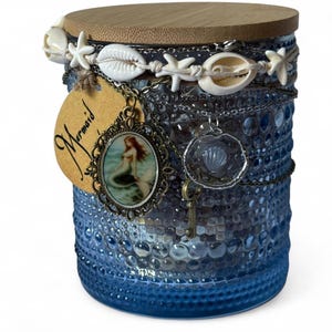 Op de afbeelding: Een blauwe glazen pot met een houten deksel, versierd met schelpen, zeesterren en kettingen. Een label met het woord "Mermaid" en een zeemeermin hanger hangen aan de pot. De pot heeft een getextureerd oppervlak.