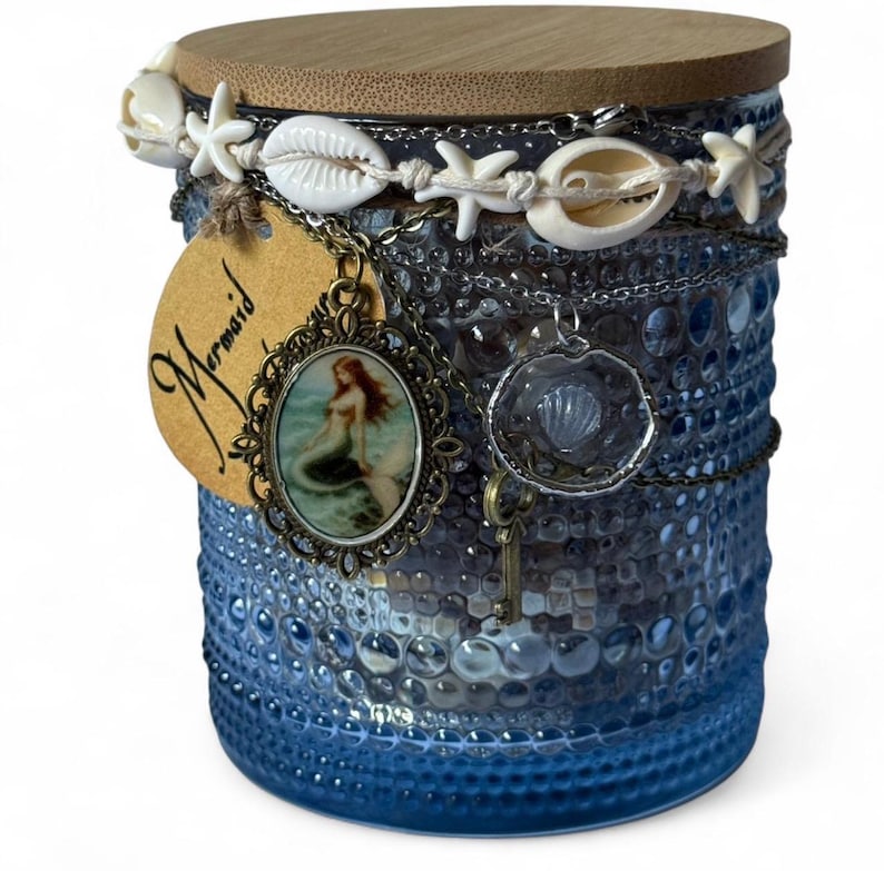 Op de afbeelding: Een blauwe glazen pot met een houten deksel, versierd met schelpen, zeesterren en kettingen. Een label met het woord "Mermaid" en een zeemeermin hanger hangen aan de pot. De pot heeft een getextureerd oppervlak.