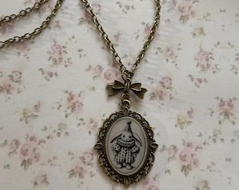 May include: A bronze chain necklace with a cameo pendant featuring a black and white illustration of a rabbit wearing a hat. The pendant is surrounded by an ornate bronze frame and is attached to the chain with a bronze bow.