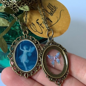 May include: Two antique bronze-toned necklaces. One pendant features a blue image of a fairy, the other a blue and pink butterfly. Both pendants are oval-shaped with ornate filigree frames and hang on delicate chains.