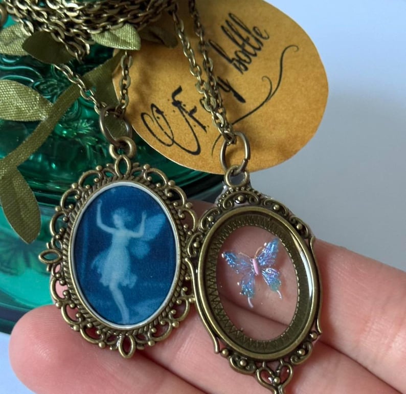 May include: Two antique bronze-toned necklaces. One pendant features a blue image of a fairy, the other a blue and pink butterfly. Both pendants are oval-shaped with ornate filigree frames and hang on delicate chains.