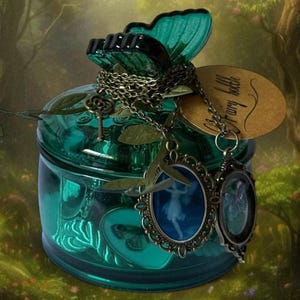 May include: A teal glass jar with a butterfly-shaped lid and decorative chain. The jar has a tag that says "Fairy bottle" and two oval pendants with fairy silhouettes. A small key charm is also attached.