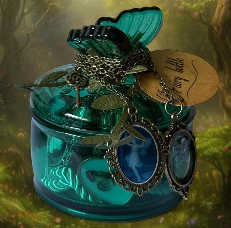 May include: A teal glass jar with a butterfly-shaped lid and decorative chain. The jar has a tag that says "Fairy bottle" and two oval pendants with fairy silhouettes. A small key charm is also attached.