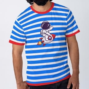 Pode incluir: Camiseta listrada azul e branca com gola e acabamento das mangas vermelhas. A camiseta apresenta um astronauta de desenho animado em um foguete vermelho e branco. O astronauta está usando um capacete e acenando. O design é centralizado na frente.