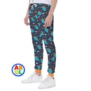 Pode incluir: Calças de moletom azul marinho com um padrão repetido de dinossauros turquesa, nuvens brancas e estrelas laranja. As calças têm punhos laranja e cintura com cordão. Tênis brancos completam o look.