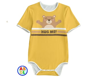 Puede incluir: Body amarillo de manga corta con un gráfico de oso marrón y el texto "Hug Me!" impreso en la parte delantera.