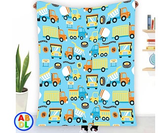 May include: A blue and yellow patterned blanket featuring a repeating design of construction trucks and cement mixers. The trucks are yellow with orange details and black wheels. The cement mixers are blue with orange details and black wheels. The background is light blue.