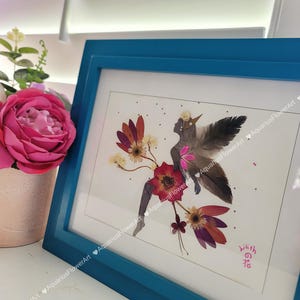 May include: A framed pressed flower art piece depicting a fairy with wings made of feathers. The fairy is surrounded by flowers in shades of pink, red, and brown. The frame is a vibrant teal blue. The text "wilith の花" is written in pink in the bottom right corner.