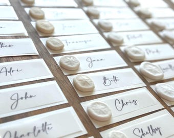 May include: Elegant place cards with names in cursive script, each adorned with a small, off-white sand dollar. The cards are arranged on a wooden surface, creating a sophisticated and beachy aesthetic.