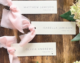 May include: Three ivory place cards with names and table numbers are displayed on a wooden surface. Each card is adorned with a delicate pink ribbon. The names are printed in a minimalist font, ideal for weddings or events.