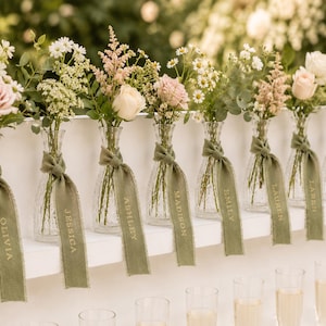May include: A row of clear, ribbed glass bud vases, each containing a small floral arrangement of pink roses, white daisies, and greenery. Each vase is tied with a sage green frayed ribbon featuring the names Olivia, Jessica, Ashley, Madison, Emily, Lauren, Karen, and Kayla.