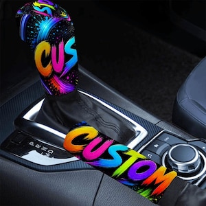 May include: A black gear shift knob cover and a matching black parking brake handle cover, both featuring a vibrant, multi-colored brushstroke design in shades of neon pink, orange, blue, and yellow, with the word CUSTOM printed in bold letters.