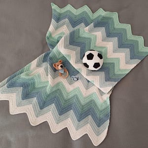 Könnte beinhalten: Eine gehäkelte Babydecke mit einem Zickzackmuster in Grün- und Blautönen. Die Decke hat einen weißen Rand und ist mit einem kleinen Stofftier und einem Fußball verziert.