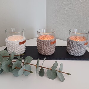 Puede incluir: Tres portavelas de vidrio con cubiertas de ganchillo. Las cubiertas son de color blanco y gris. Los portavelas están sobre una superficie de pizarra negra.