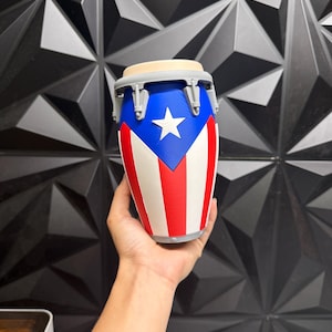 May include: A small, decorative conga drum featuring the flag of Puerto Rico. The drum has a blue top with a white star, and red and white stripes on the body. The drum is held in a person's hand.