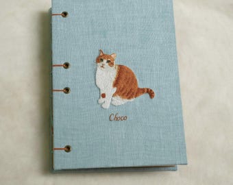 Puede incluir: Un diario de lino azul claro con un gato marrón bordado en la parte delantera. El gato es blanco y naranja con ojos marrones. El nombre del gato, "Choco", está bordado debajo del gato.