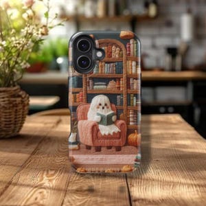 May include: A phone case with a charming design of a ghost reading a book in an armchair. The background shows a detailed bookshelf with various books. The case has a dark background with a Halloween theme.