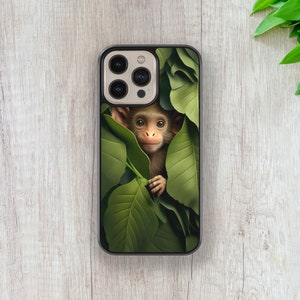 May include: A phone case featuring a cute monkey peeking out from behind green leaves. The monkey has a light brown face and a white nose.