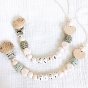 Può includere: Due morsi per bambini in legno con clip, ognuno con un nome diverso scritto in perle di legno. I nomi sono "Kais" e "Leon". I morsi hanno anche cuori di legno e perline di uncinetto verdi.