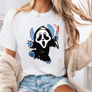 May include: White t-shirt with a cartoon character mashup of Stitch and the Ghostface mask from the Scream films. The character is black and white with blue accents, holding a bloody knife. The design is centered on the shirt.