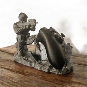 May include: A gray 3D printed figurine of a soldier holding a gun, kneeling on a gray, rocky base. The soldier is holding a black video game controller.