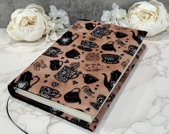 May include: A hardback book with a brown cover featuring a repeating pattern of coffee-related illustrations in black. The illustrations include coffee cups, teapots, and text. A black ribbon bookmark is visible. The background includes white flowers.