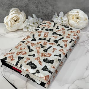 May include: A book cover featuring a repeating pattern of black and brown chess pieces on a cream background. The cover has a black spine with a red accent and a black ribbon bookmark. White flowers are in the background.