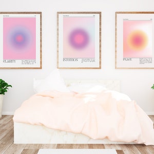 May include: Three framed prints with abstract gradient designs in pink, purple, and yellow. The prints are titled "Clarity", "Intuition", and "Peace" and are displayed on a white wall above a bed with a white headboard and a pink duvet.