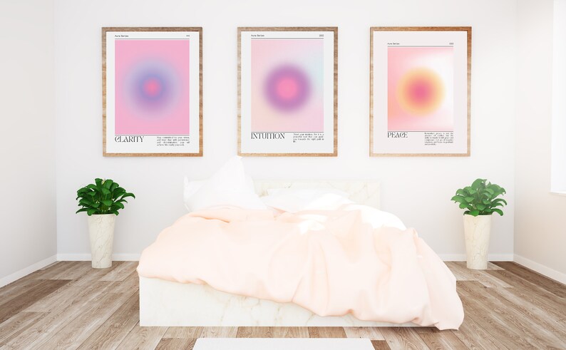 May include: Three framed prints with abstract gradient designs in pink, purple, and yellow. The prints are titled "Clarity", "Intuition", and "Peace" and are displayed on a white wall above a bed with a white headboard and a pink duvet.