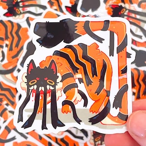 May include: A sticker depicting a cartoon creature with a black cat-like head, an orange body, and black stripes. The creature has long black tentacles. Similar stickers are in the background.