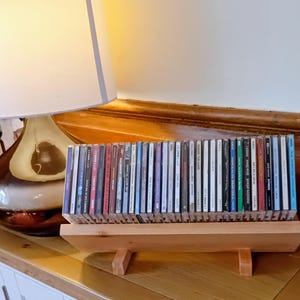 Puede incluir: Un estante de madera para CD que contiene una colección de CD. El estante está hecho de madera clara y tiene un diseño simple y minimalista. Los CD están dispuestos en una fila, con sus lomos hacia adelante. Los lomos muestran varios colores y texto.