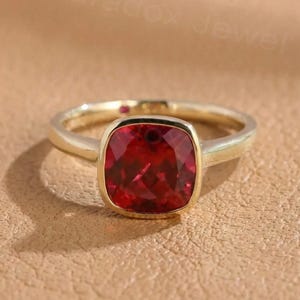 Puede incluir: Un anillo de oro con una gran piedra preciosa roja oscura de talla cuadrada. La piedra preciosa está engastada en un bisel de oro, y la banda es de oro pulido simple. El anillo se muestra sobre una superficie texturizada de color canela.