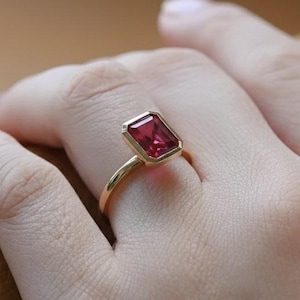 Peut inclure: Une bague en or ornée d'une pierre précieuse rectangulaire rouge foncé. La bague a un fin anneau poli, et la pierre est sertie dans un sertissage à lunette. La bague est présentée sur une main, mettant en valeur son design élégant.