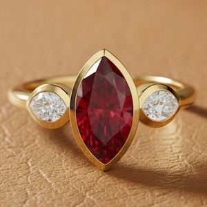 以下が含まれることがあります： 中央に大きなマーキス型の深い赤色の宝石、両側に2つの小さな丸い透明な宝石をあしらったゴールドリング。 リングは、ジュエリーのデザインと職人技を際立たせる、質感のあるタン色の表面にセットされています。