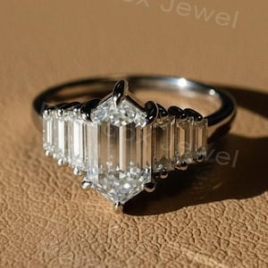 Op de afbeelding: Een zilveren ring met heldere, smaragdgeslepen stenen. De centrale steen is zeshoekig, geflankeerd door kleinere rechthoekige stenen. De ring ligt op een getextureerd, bruin oppervlak. De tekst "Paradox Jewel" is zichtbaar.