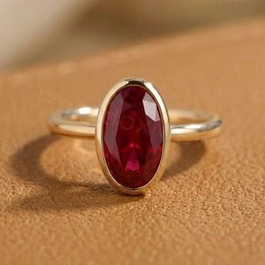 Puede incluir: Un anillo de oro con una piedra preciosa roja oscura de corte ovalado. La piedra preciosa está engastada en un engaste de bisel, y la banda es de oro pulido. El anillo se muestra sobre una superficie marrón.