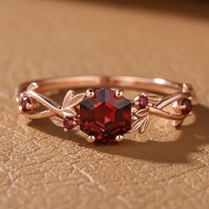 May include: Rose gold ring featuring a large, faceted, hexagonal red gemstone as the centerpiece. The band is adorned with delicate leaf and small red gemstone accents, creating an elegant, nature-inspired design.