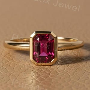 May include: A gold ring with a rectangular, deep pink gemstone. The gemstone is set in a bezel setting, and the ring band is a simple, polished gold. The background is a textured, tan surface. The text "Paradox Jewel" is visible.