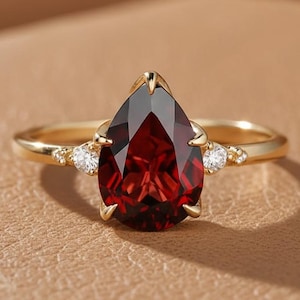 May include: A gold ring with a teardrop-shaped, deep red gemstone as the focal point. The ring is embellished with small, round, white gemstones. The band is a simple, polished gold. The ring is set against a neutral background.