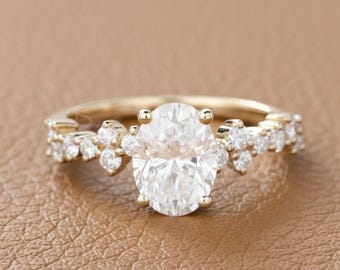 May include: A gold engagement ring with an oval-cut diamond center stone. The band features smaller accent diamonds and floral-inspired detailing. The ring is set against a brown background.