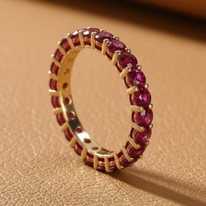 May include: A gold-colored ring with a continuous band of small, round, red gemstones. The ring is set against a neutral, textured background, highlighting the vibrant color of the stones and the ring's design.