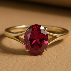 Puede incluir: Un anillo de oro con un gran rubí de corte ovalado. El rubí es de un color rojo intenso y está engastado en una montura de cuatro garras. La banda del anillo tiene un diseño dividido, añadiendo un toque moderno.