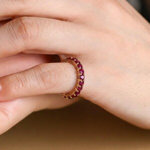 May include: A gold ring set with a continuous line of small, round, red gemstones. The ring is on a finger, with the hand and part of the arm in view. The ring's design is simple, with the gemstones closely set.